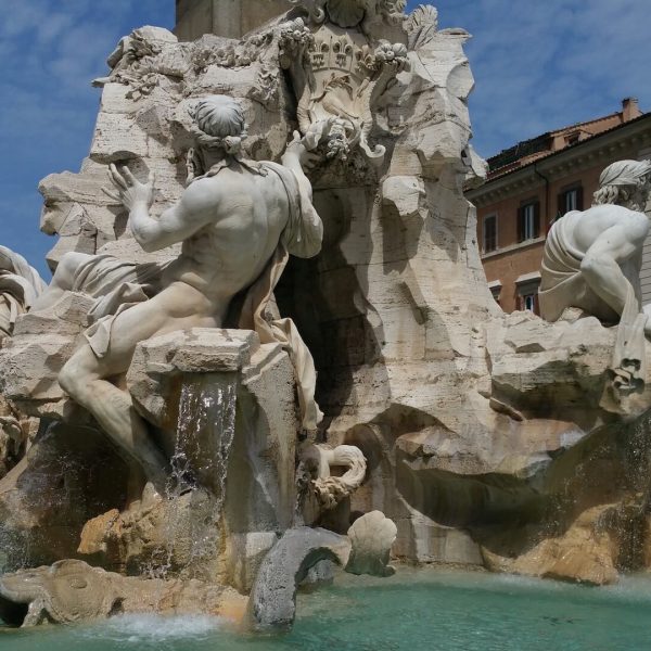 Ornate stone fountain with multiple sculpted figures and water flowing into a turquoise basin, situated in a square with surrounding historic buildings under a clear blue sky.