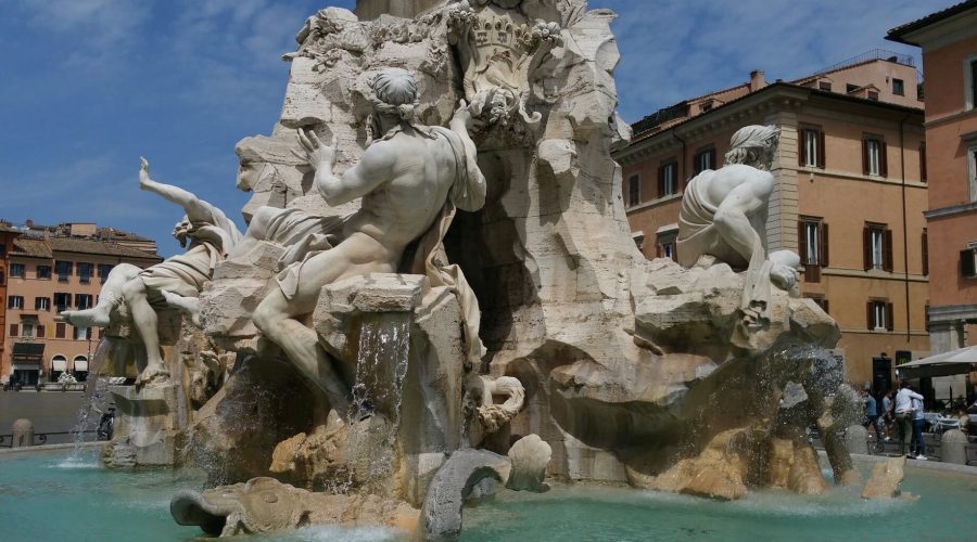 Ornate stone fountain with multiple sculpted figures and water flowing into a turquoise basin, situated in a square with surrounding historic buildings under a clear blue sky.