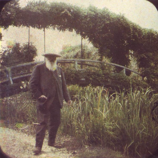 An elderly man with a long beard and broad-brimmed hat stands in a garden in front of an arched bridge covered with foliage.