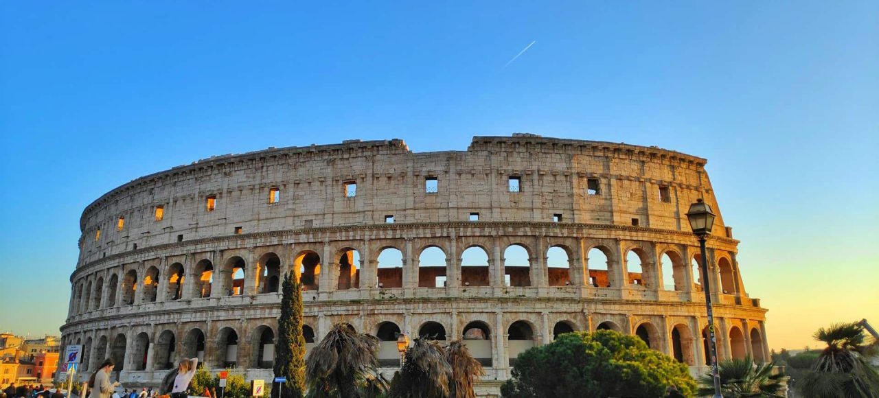 A wide view of the Colosseum in Rome, Italy, during sunset. The ancient amphitheater is partially illuminated by the setting sun, surrounded by trees and a few people in the foreground.