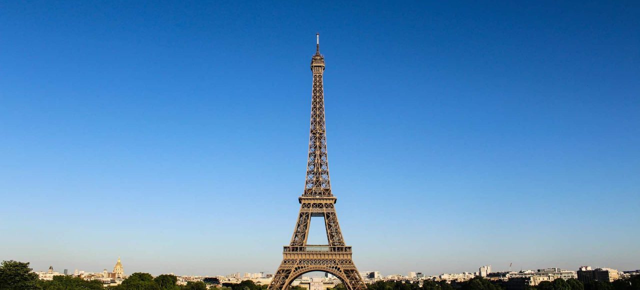 The Eiffel Tower in Paris stands tall against a clear blue sky, surrounded by greenery and buildings in the distance.